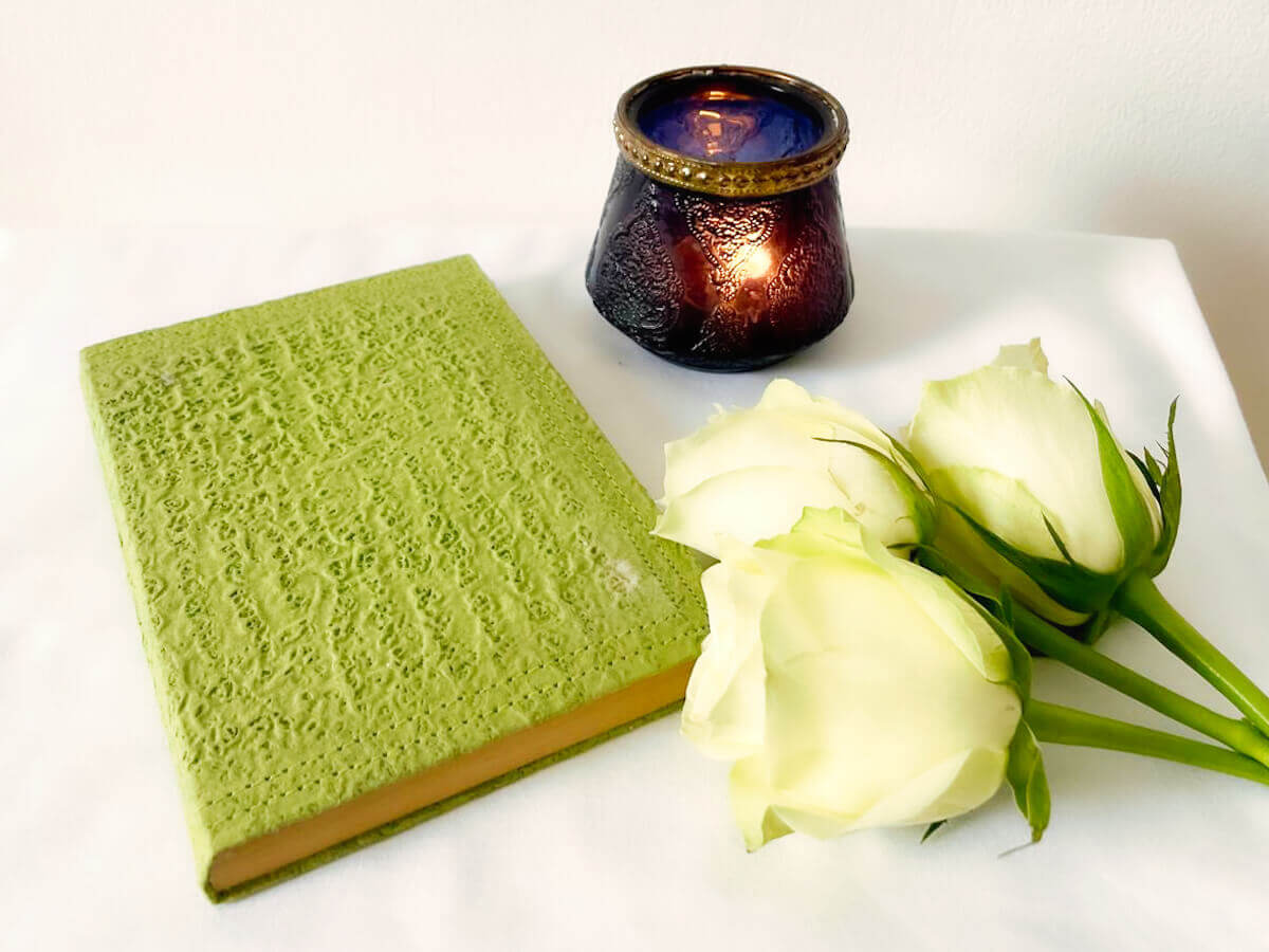 A hardcover healing book rests on a table with a lit candle and white roses. It is used to send out healing energy, especially at 10 pm UK time, a common time for absent healing.